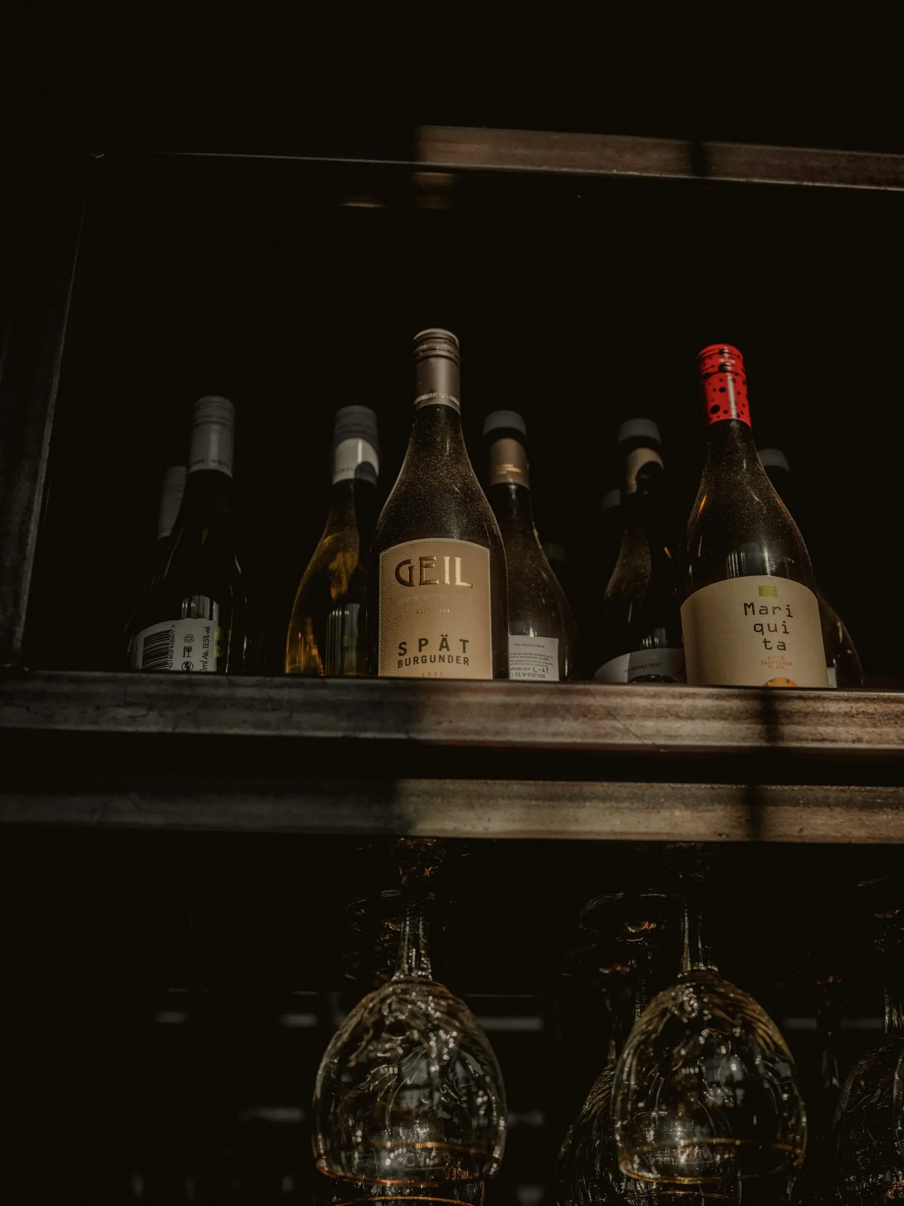 Natural wine bottles on a dark wooden shelf above hanging glasses