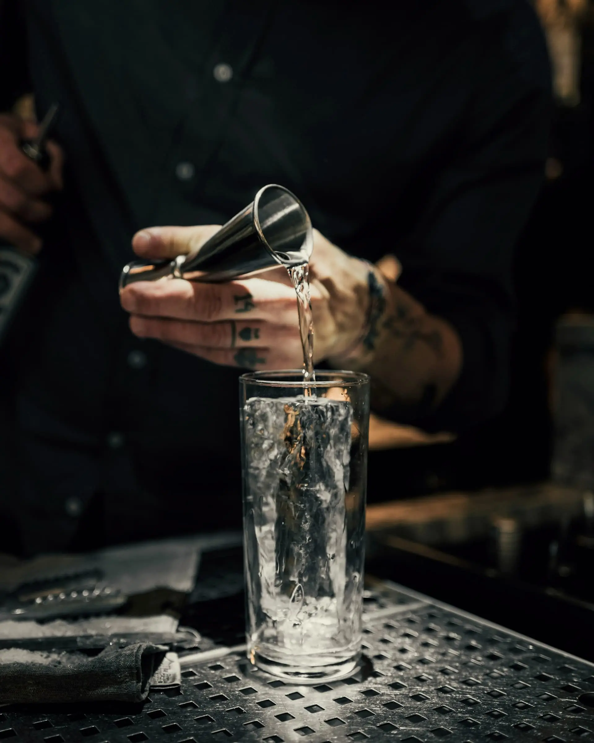 A tattooed hand pouring from a steel jigger into a mixing glass