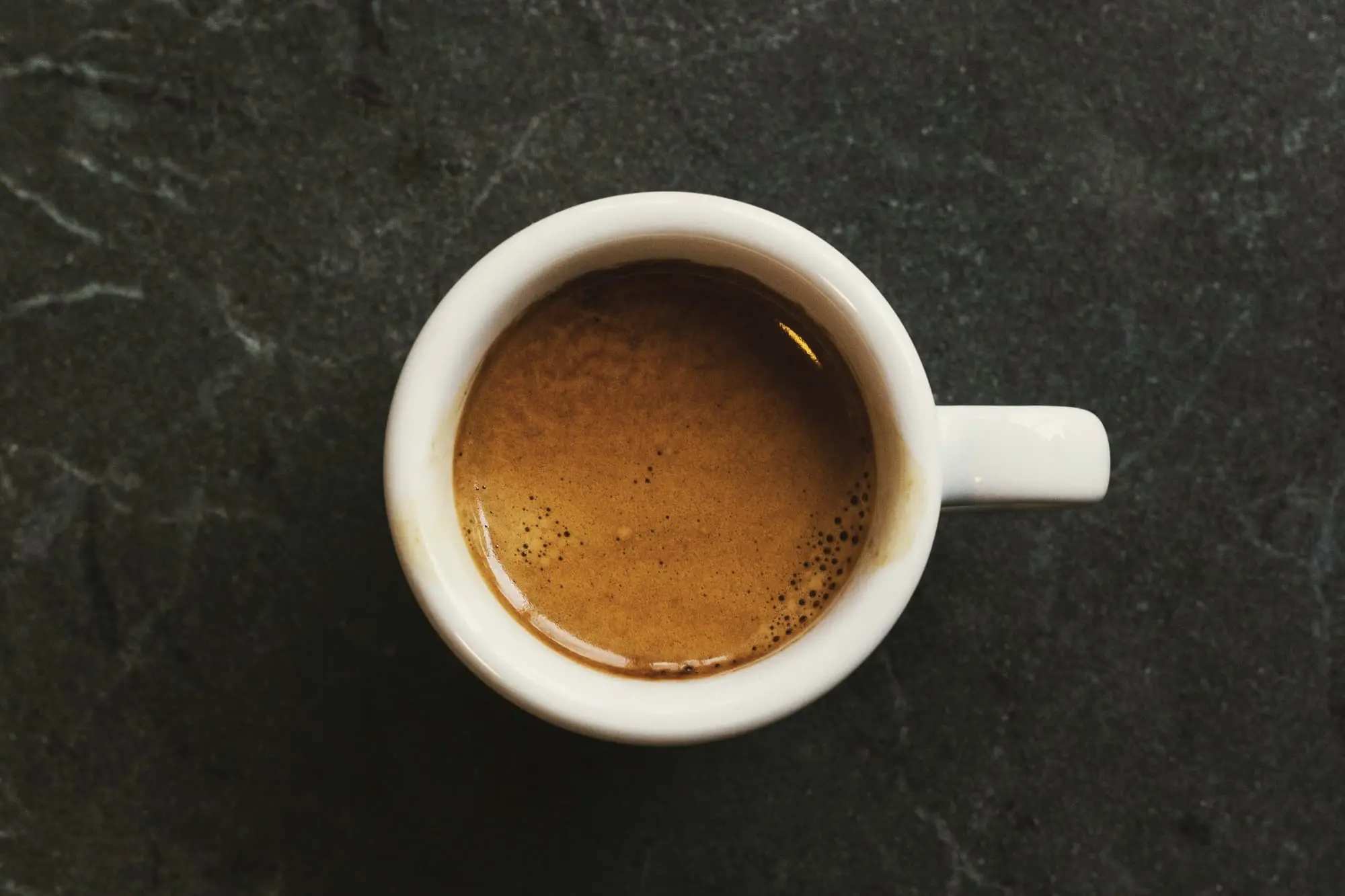 A white cup of espresso seen from above on black stone