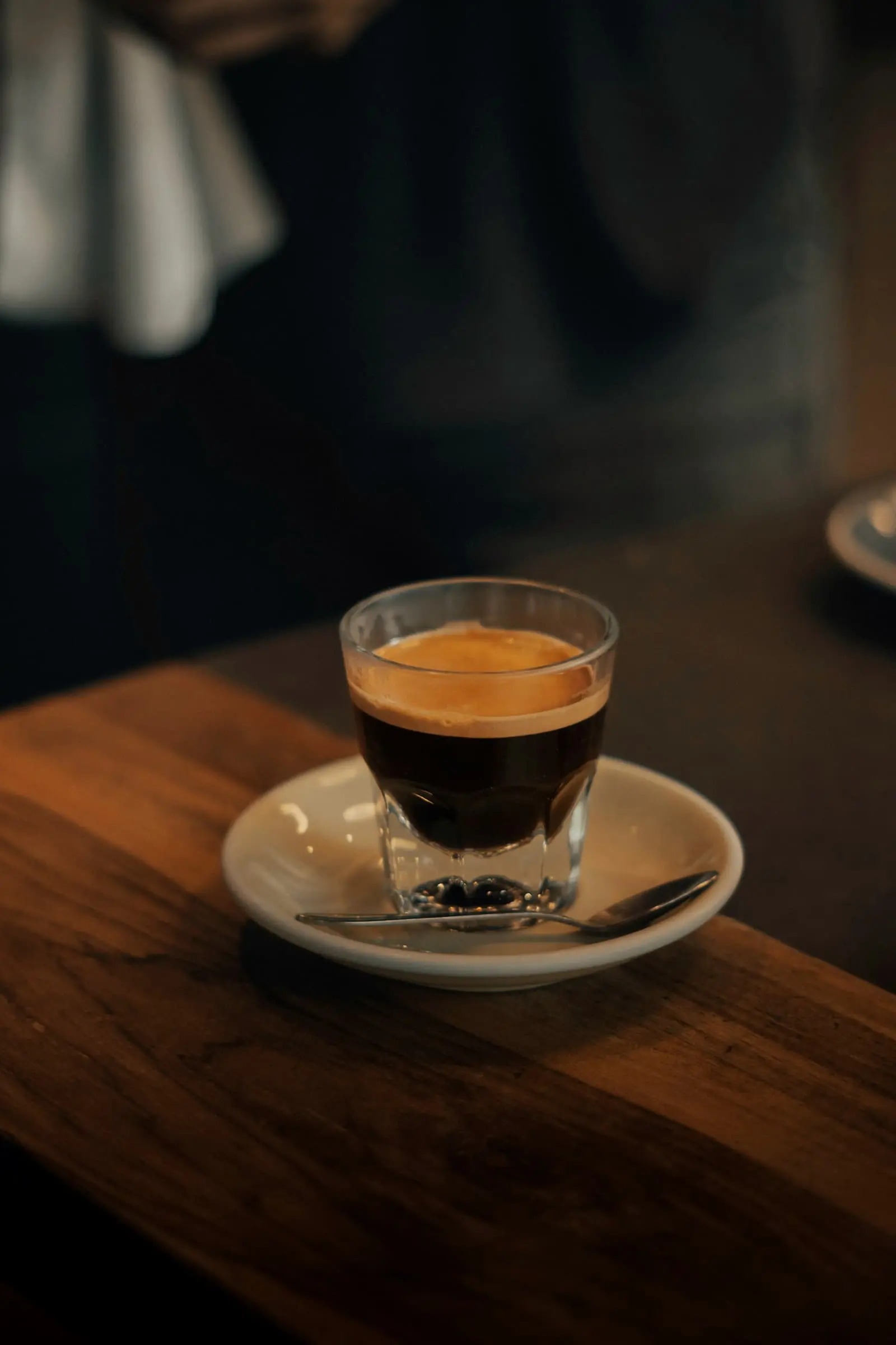 A short coffee in a glass on a saucer at the edge of a wooden table
