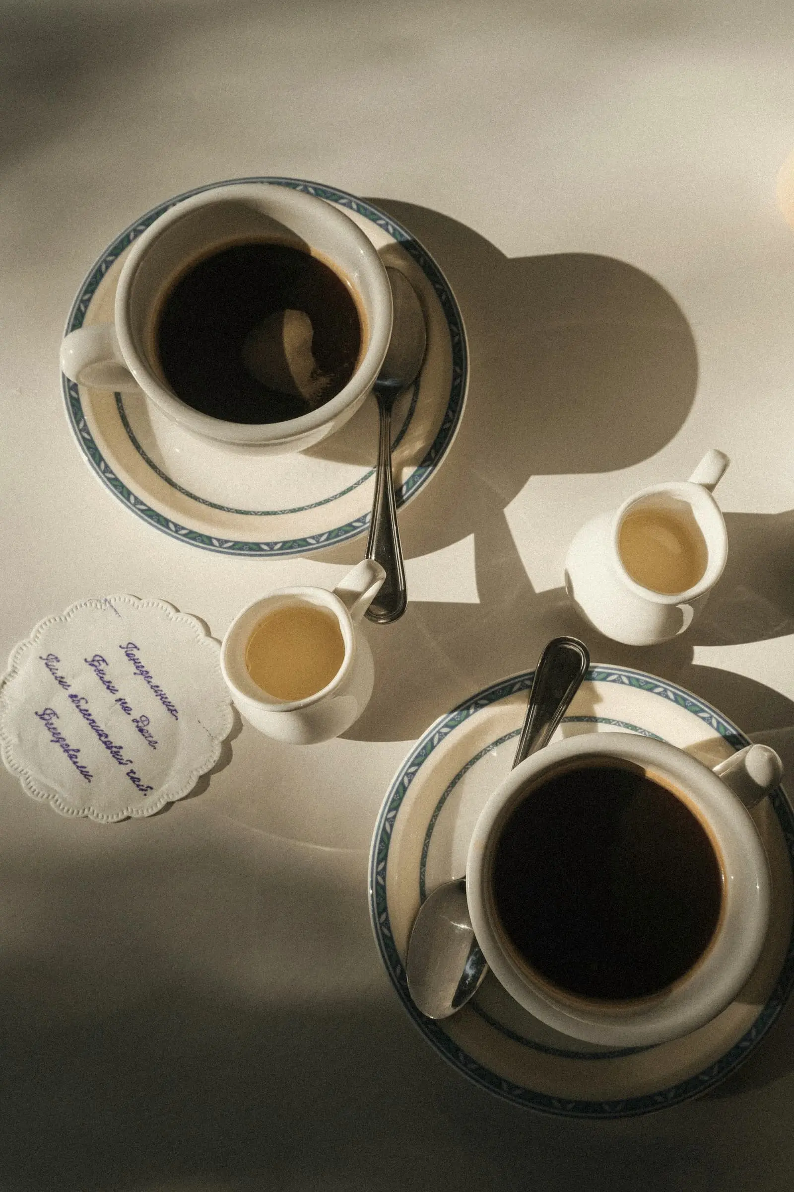 Two cups of black coffee and two small milk jugs on a sunlit table, seen from above