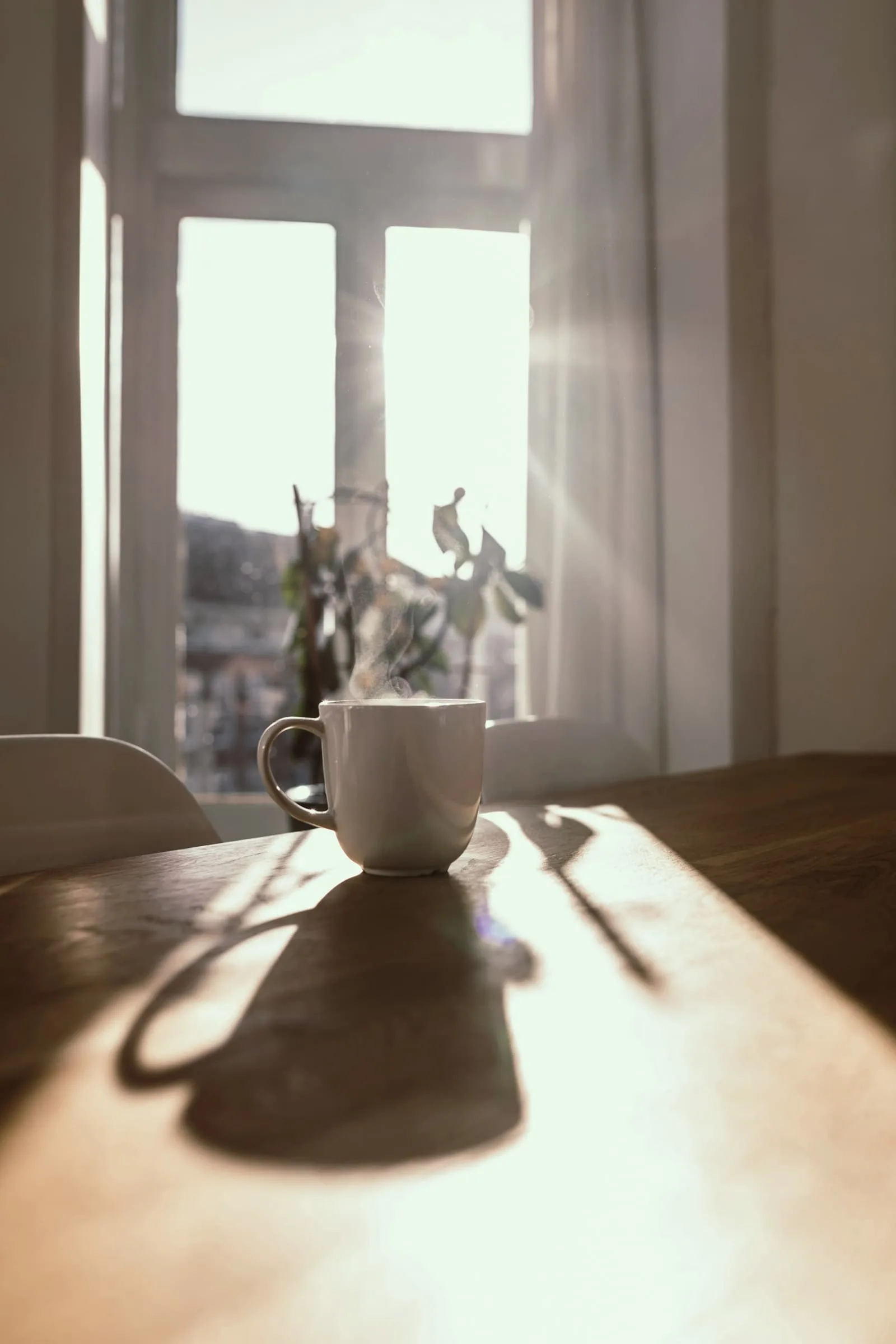 Steam rising from a mug on a wooden table before a bright curtained window