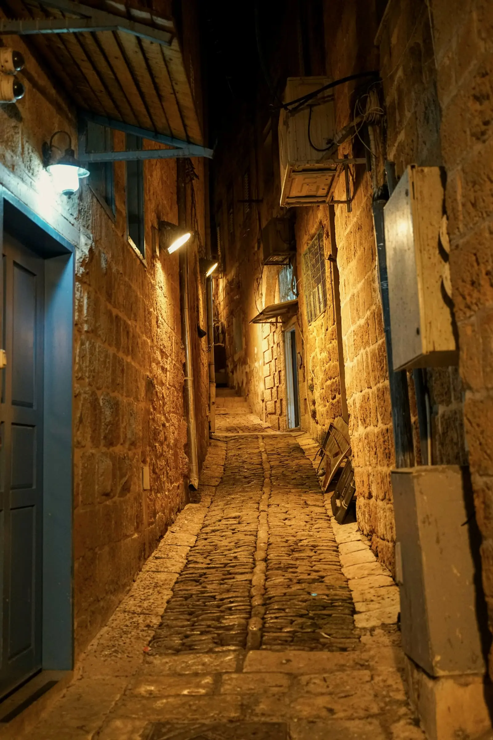 A narrow stone lane at night, its brick walls amber under the wall lamps