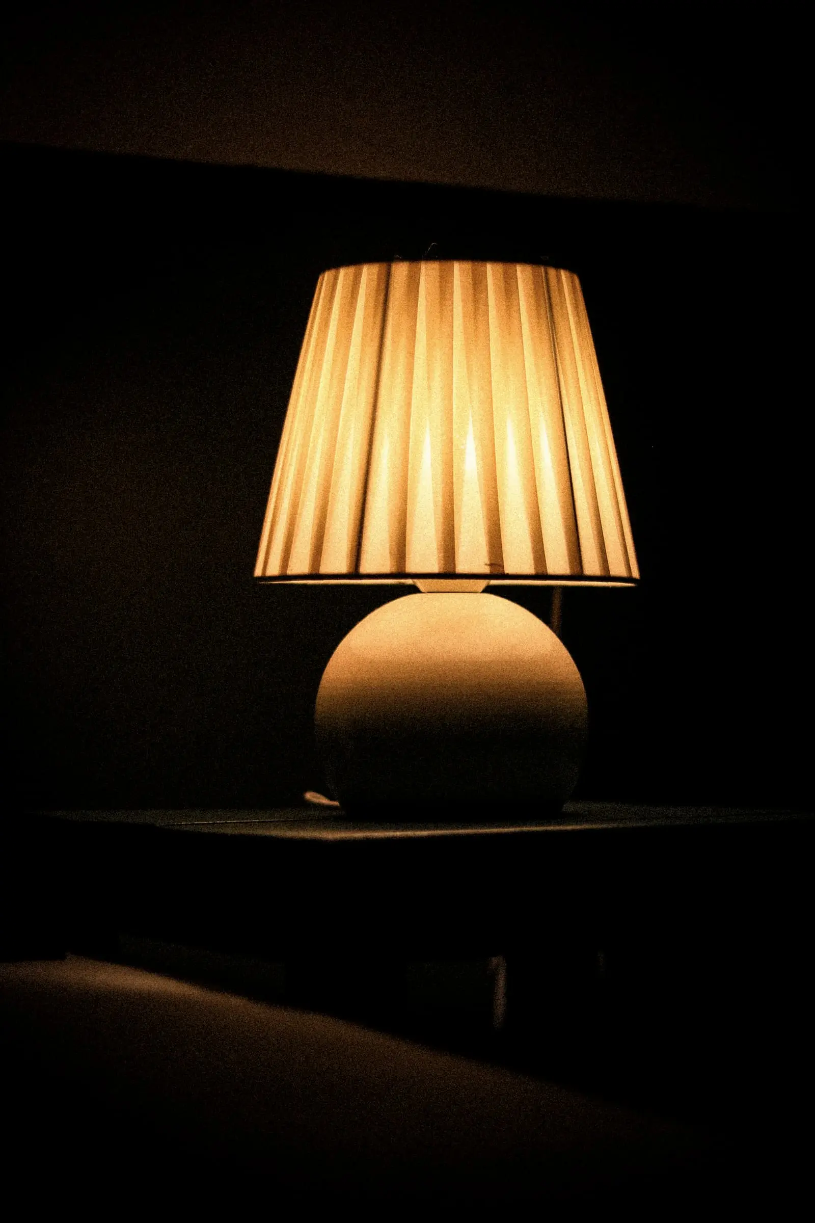A pleated lampshade glowing amber on a dark table