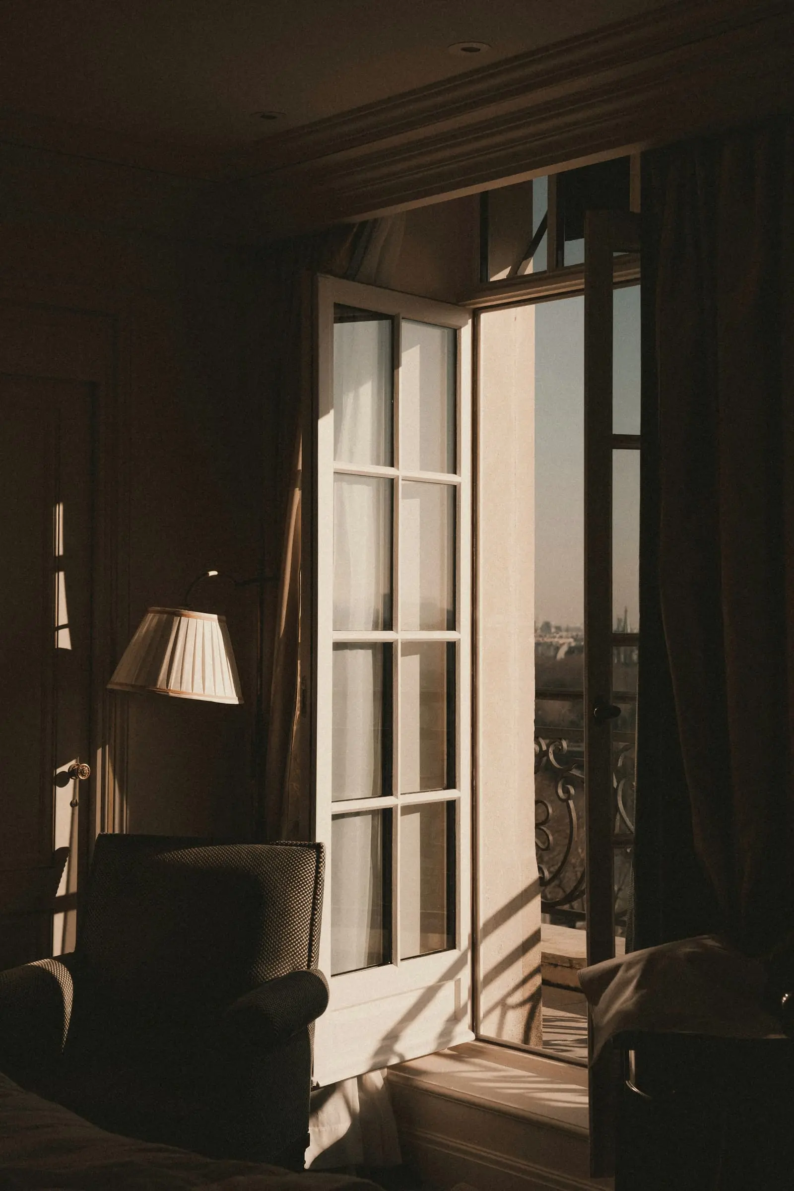 Open French windows in a dark room with a lamp and armchair beside them