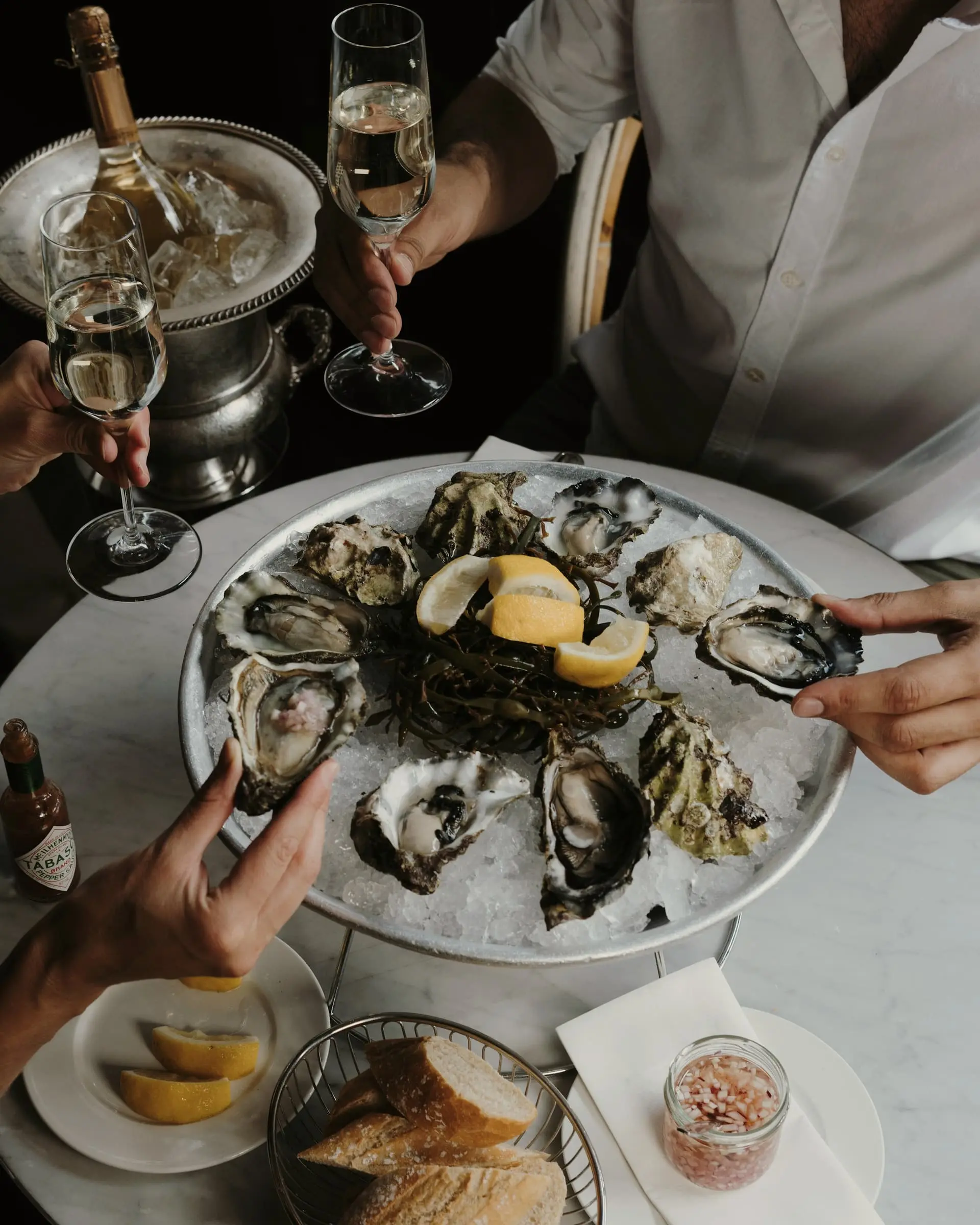 Hands reaching for oysters and lemon on a platter, sparkling wine alongside