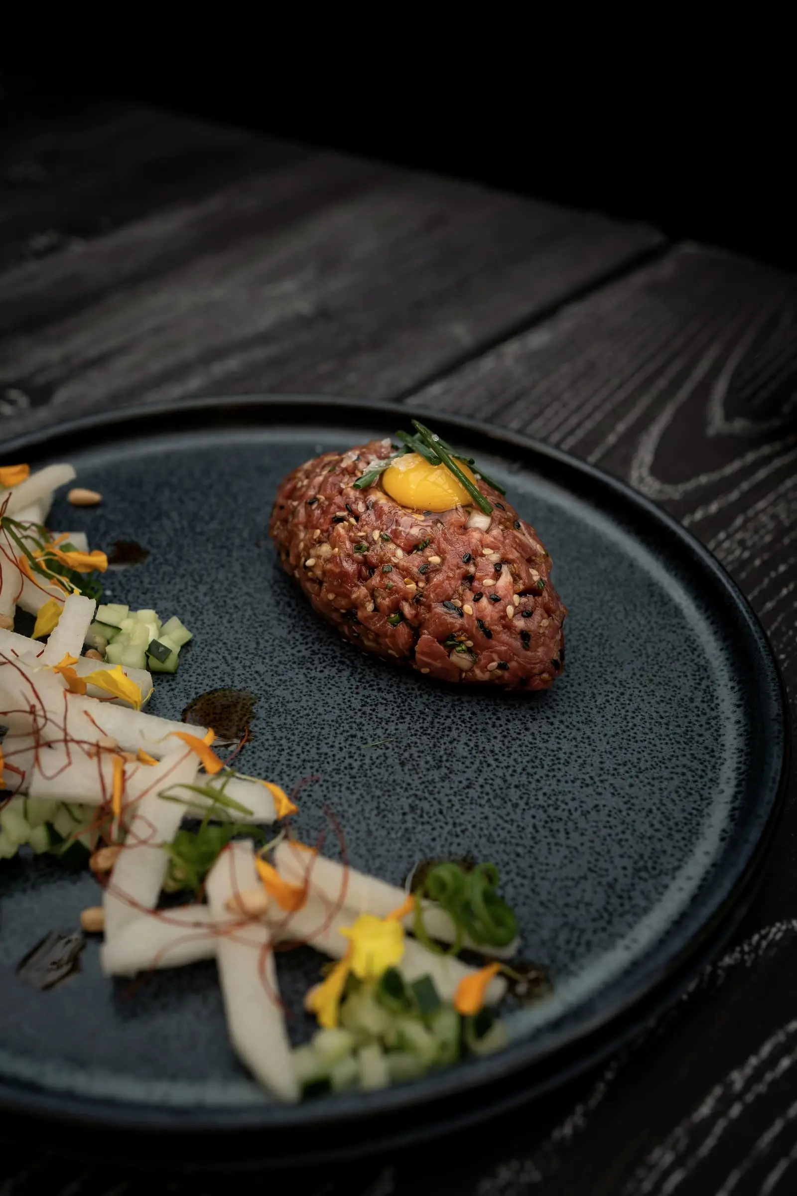 Hand-cut beef tartare with a yolk on a dark plate, pickles alongside