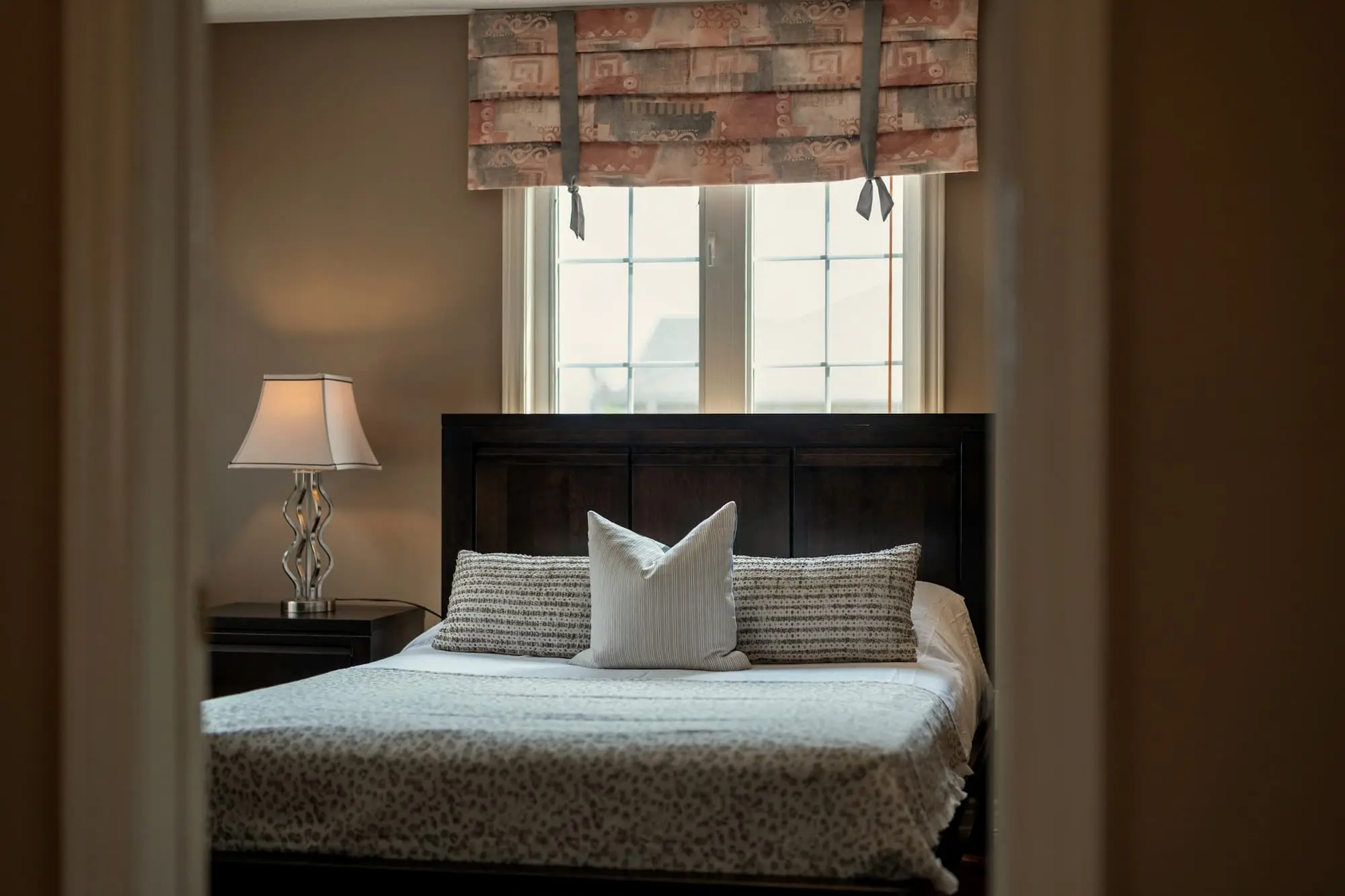Bedroom seen through a doorway, a dark bed beneath a window with a Roman blind
