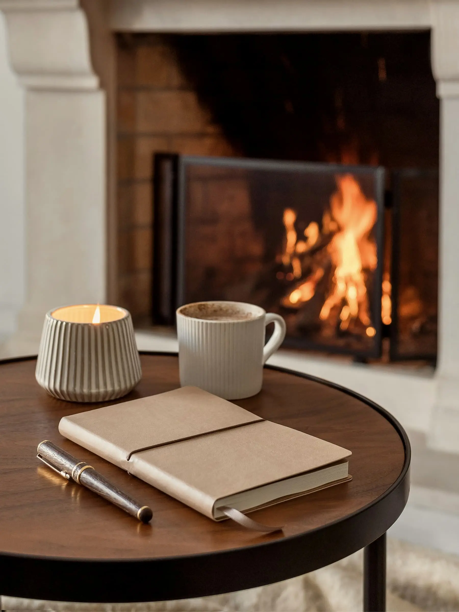 A candle, a mug and a notebook on a round table before a lit fire