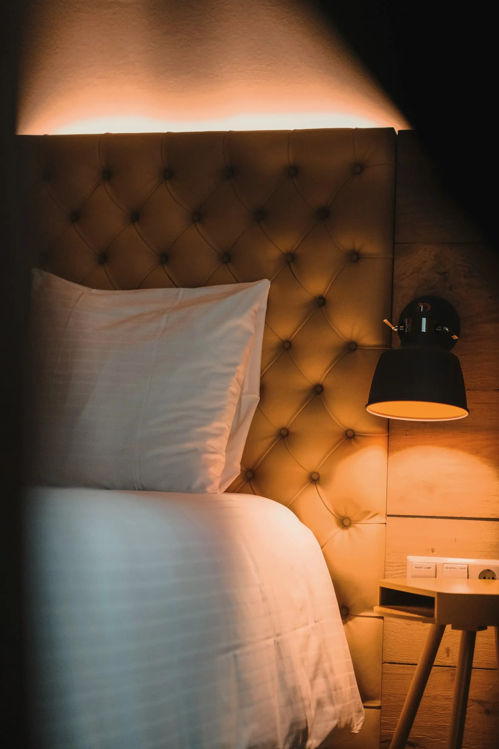 A deep-buttoned amber headboard lit by a black wall lamp, white pillows against it