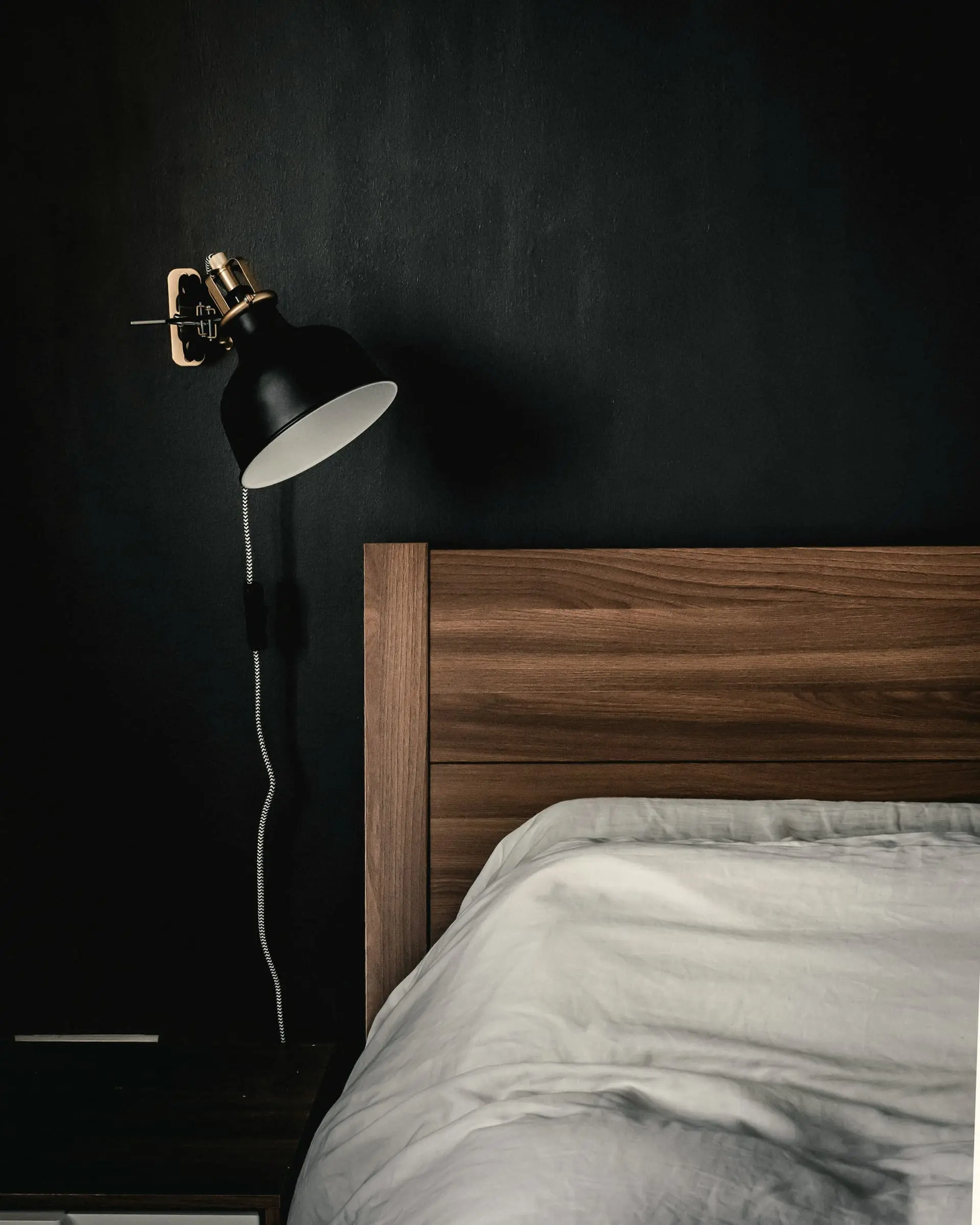 A walnut bed end and black wall lamp against a dark green wall