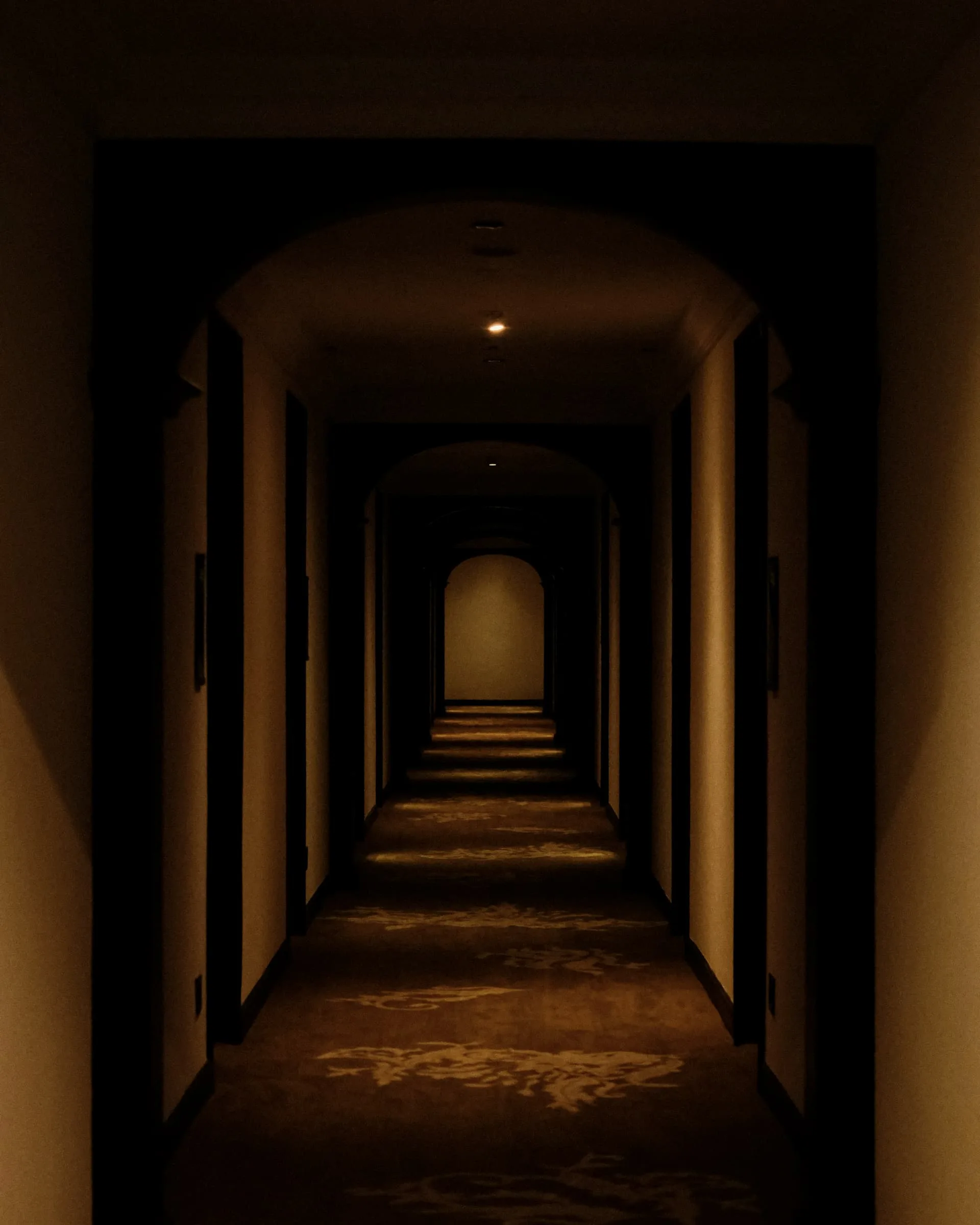 A dim corridor of receding arches, warm pools of light along a patterned carpet