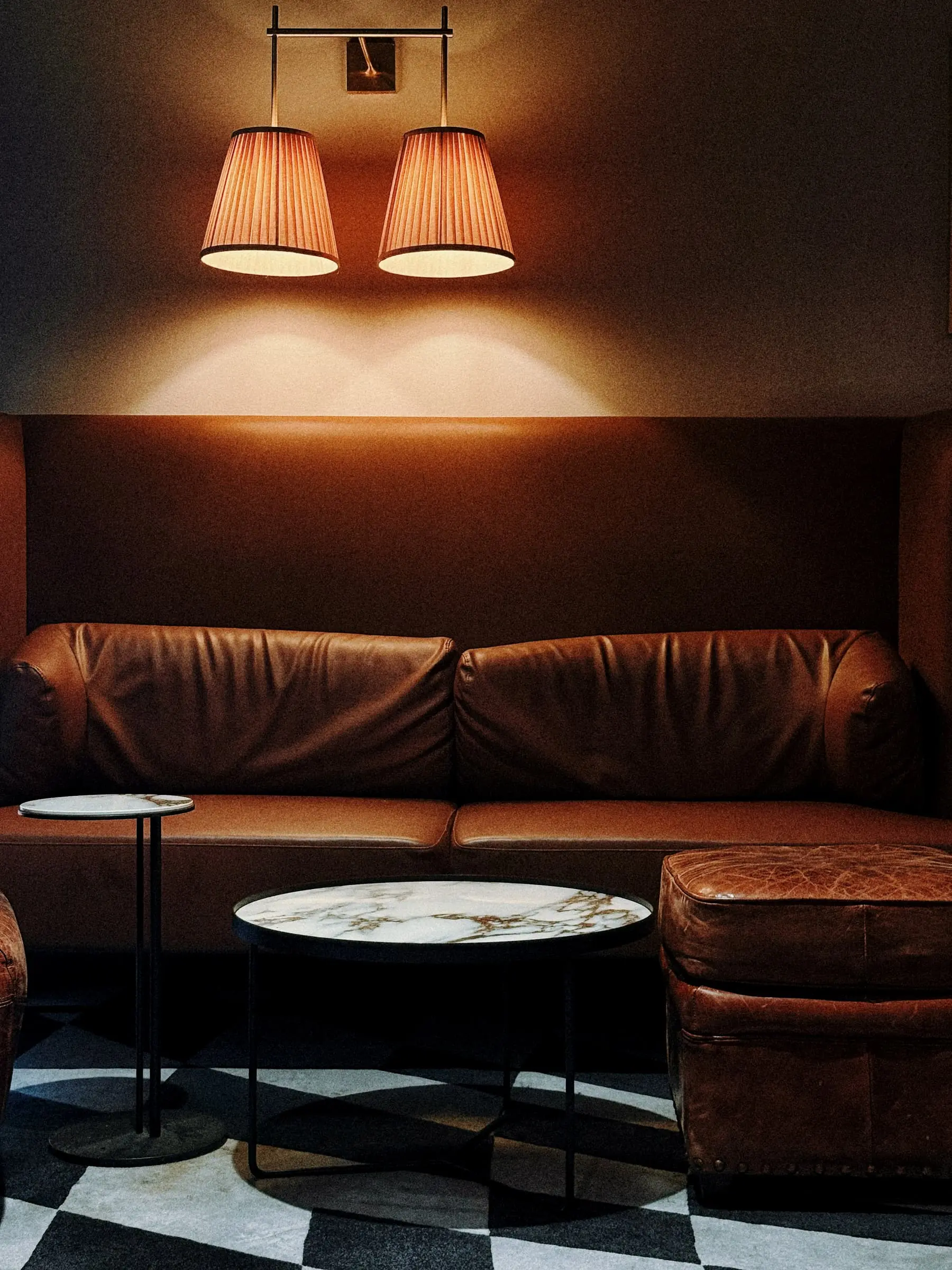 A tan leather banquette and marble table under twin lamps, on a checked floor