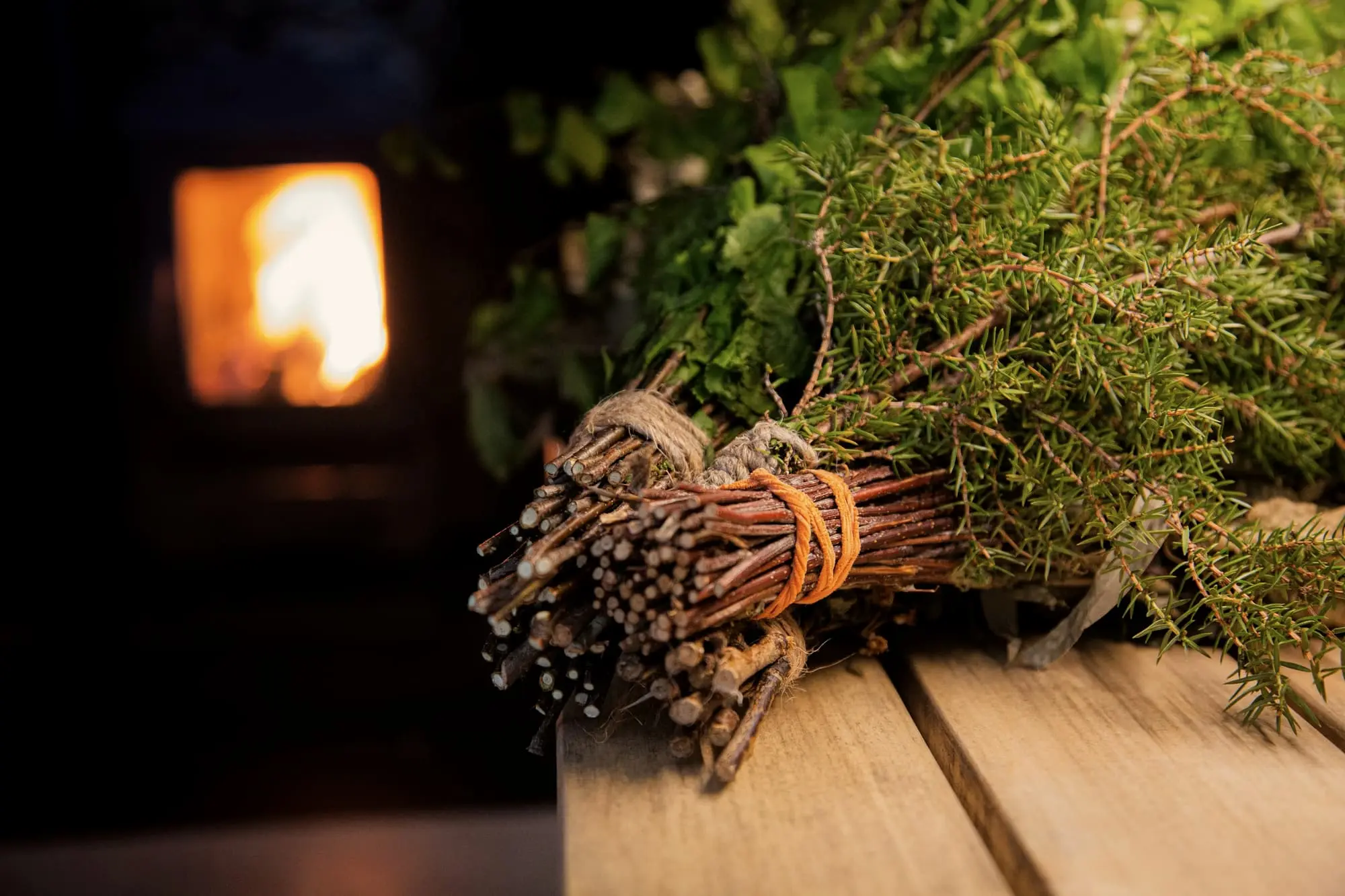A bundle of birch and herbs on a sauna bench, stove glowing behind